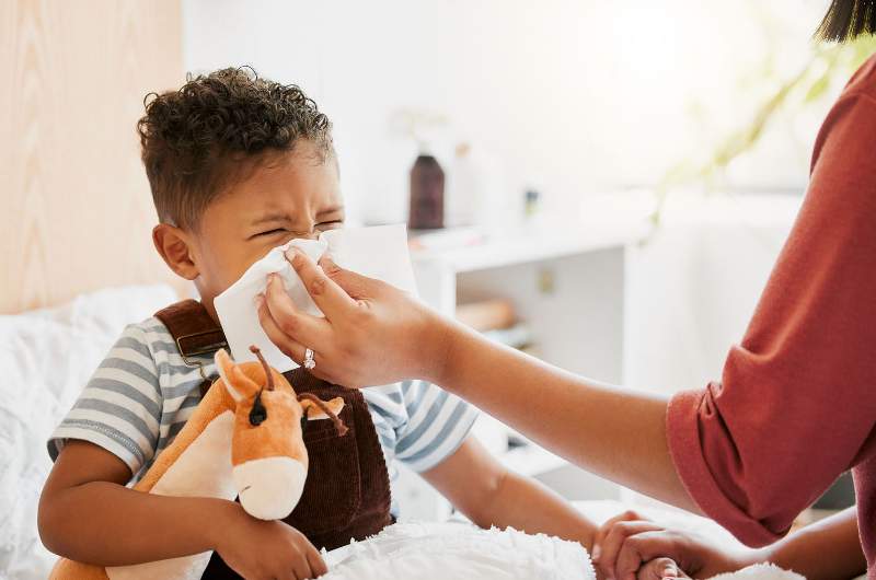 An unwell little boy with the flu holds his toy giraffe while his mother helps to wipe his nose.