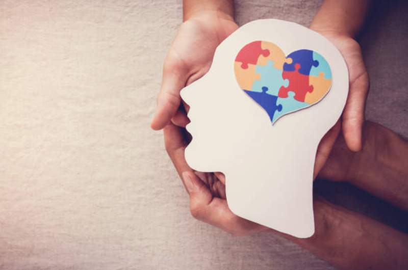 Wooden human head outline with puzzle pieces inside head in heart shape held by two sets of hands depicting mental health