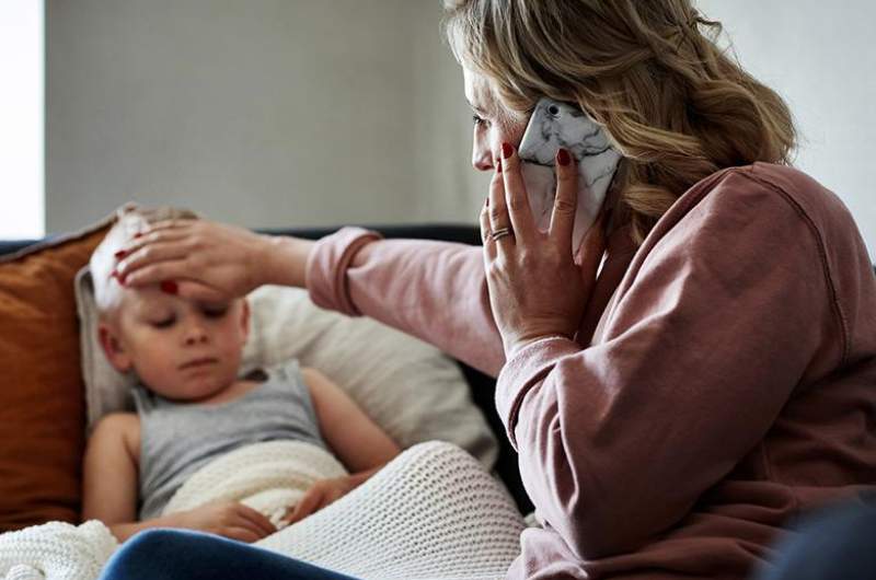 Mother on phone with telehealth doctor appointment on a Sunday, with hand held up to unwell child's forehead.