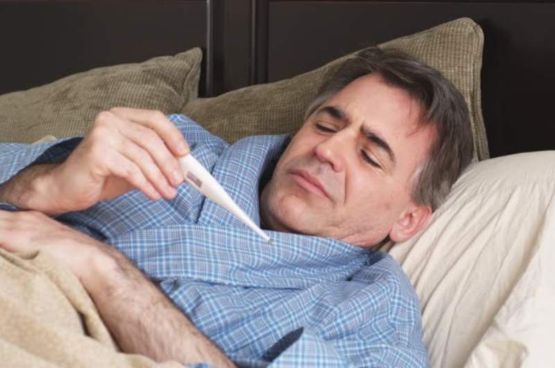 Person lying in bed ill with yellow fever looking at thermometer experiencing fever.