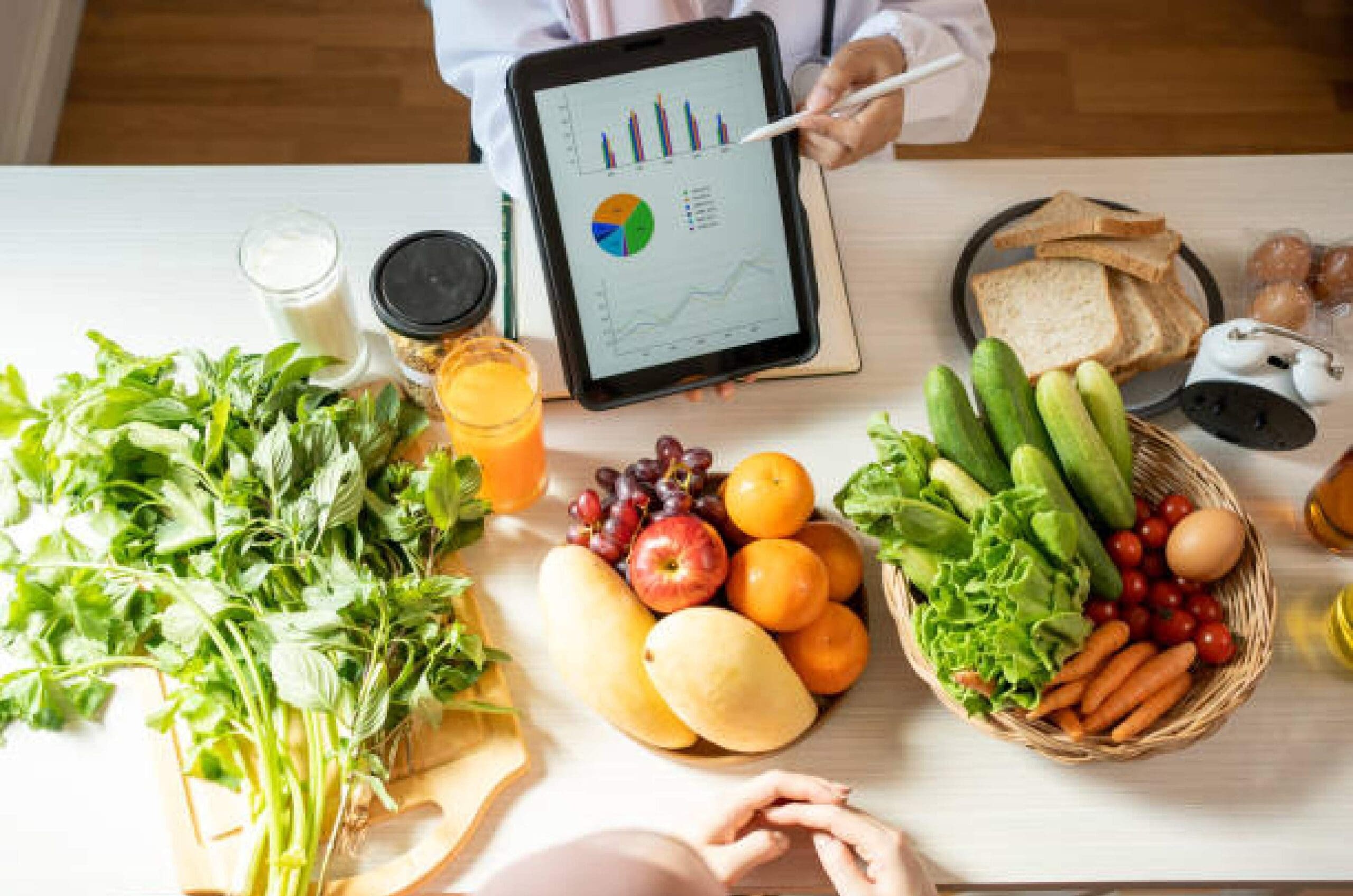 Nutritionist-table-with-vegetables-whole-foods-and-information Nutritionist's desk showcasing whole foods, vegetables and fruits, while displaying infographic on tablet screen, sharing nutritional information and education for patient.