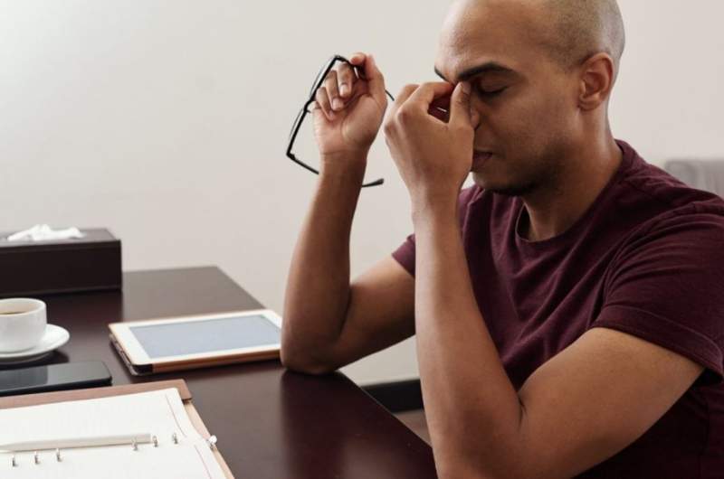 A man sits at a desk with eyes closed, while pinching his upper nose and appearing tired with low energy.