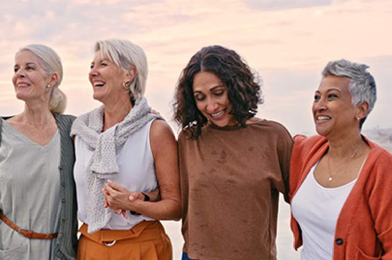 Four older women of various backgrounds who are experiencing or have experienced menopause, stand together with arms around each other with a beautiful sunrise sky behind them.