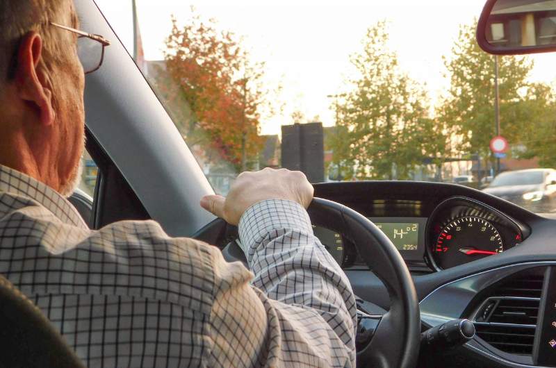 An elderly man aged over 75 years old, is in the driving seat of a car with his hands on the steering wheel as his car stops at a red traffic light.