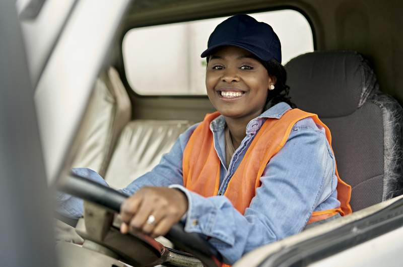 A young female commercial truck driver sitting in the cabin of her truck smiling with her hands on the steering wheel.